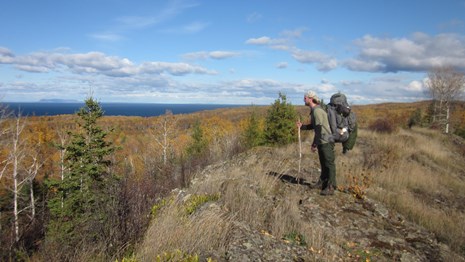 A person with a backpack stands on a rocky ridge looking over a forest and a lake. 