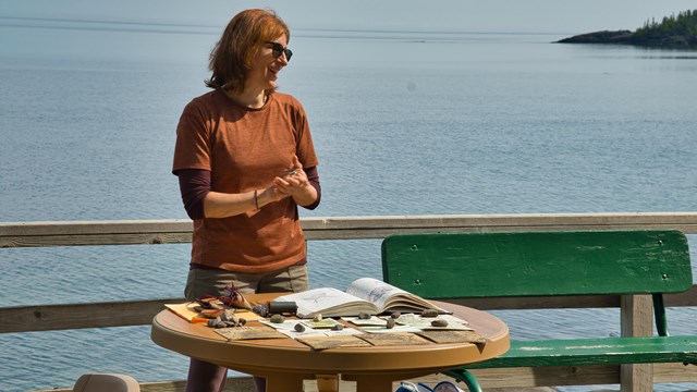 A smiling woman in an orange shirt stands at a round table displaying artwork.