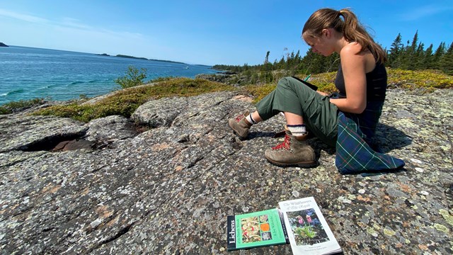 Teen Artist sitting on rocky Lake Superior shoreline, sketching with colored pencils.
