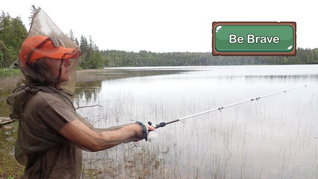 A person wearing mosquito netting fishing on a grassy lake.
