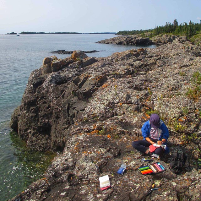 Teen Artist sitting on rocky Lake Superior shoreline, sketching with colored pencils.