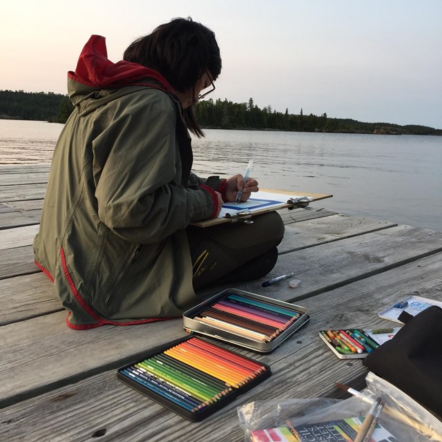 A young woman sits on a dock over a lake and draws with art supplies around her