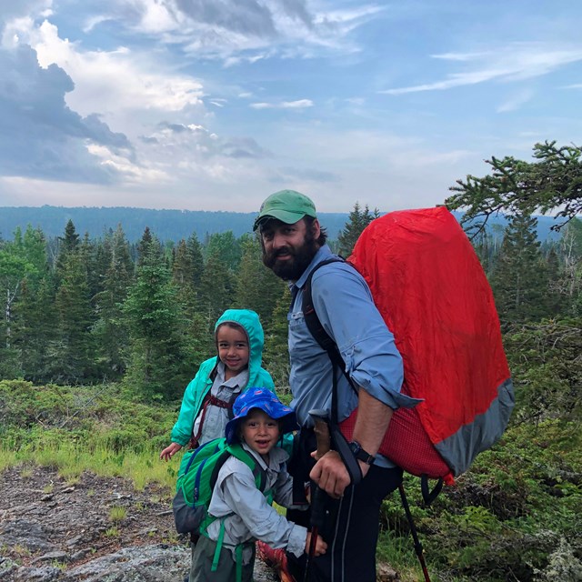 An adult with a backpack on stands next to two children along a rocky trail in a forest.
