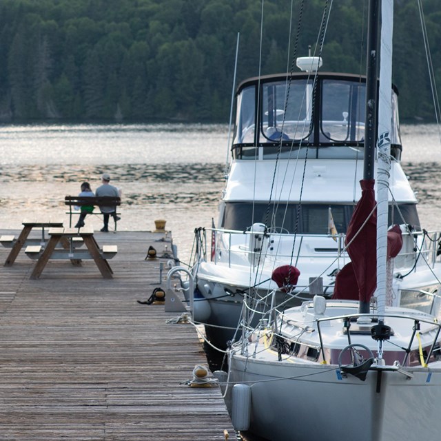 Boats tied up at a dock and two people sitting on a bench at the end of the dock.