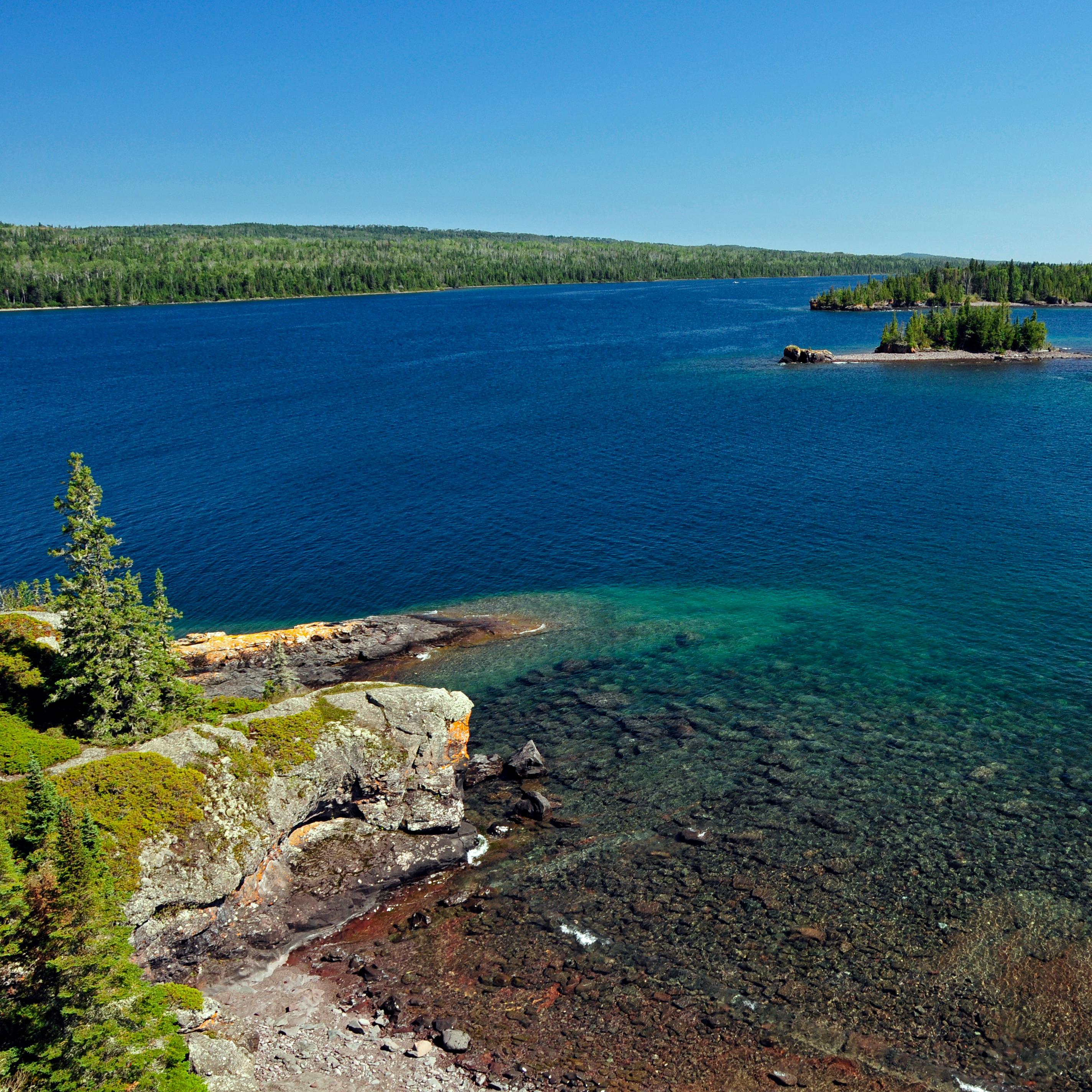 Rugged Isle Royale coastline with blue water, shallow reefs, forest, and rocks.