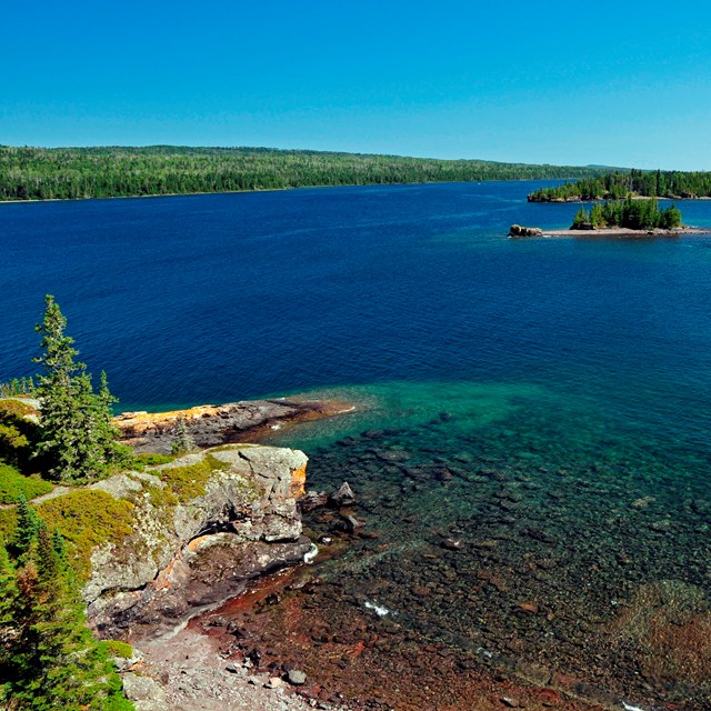 Rugged Isle Royale coastline with blue water, shallow reefs, forest, and rocks.