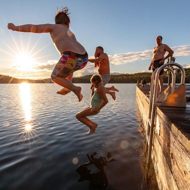 Two people jump off of a dock into calm waters as the sun sets in the background.