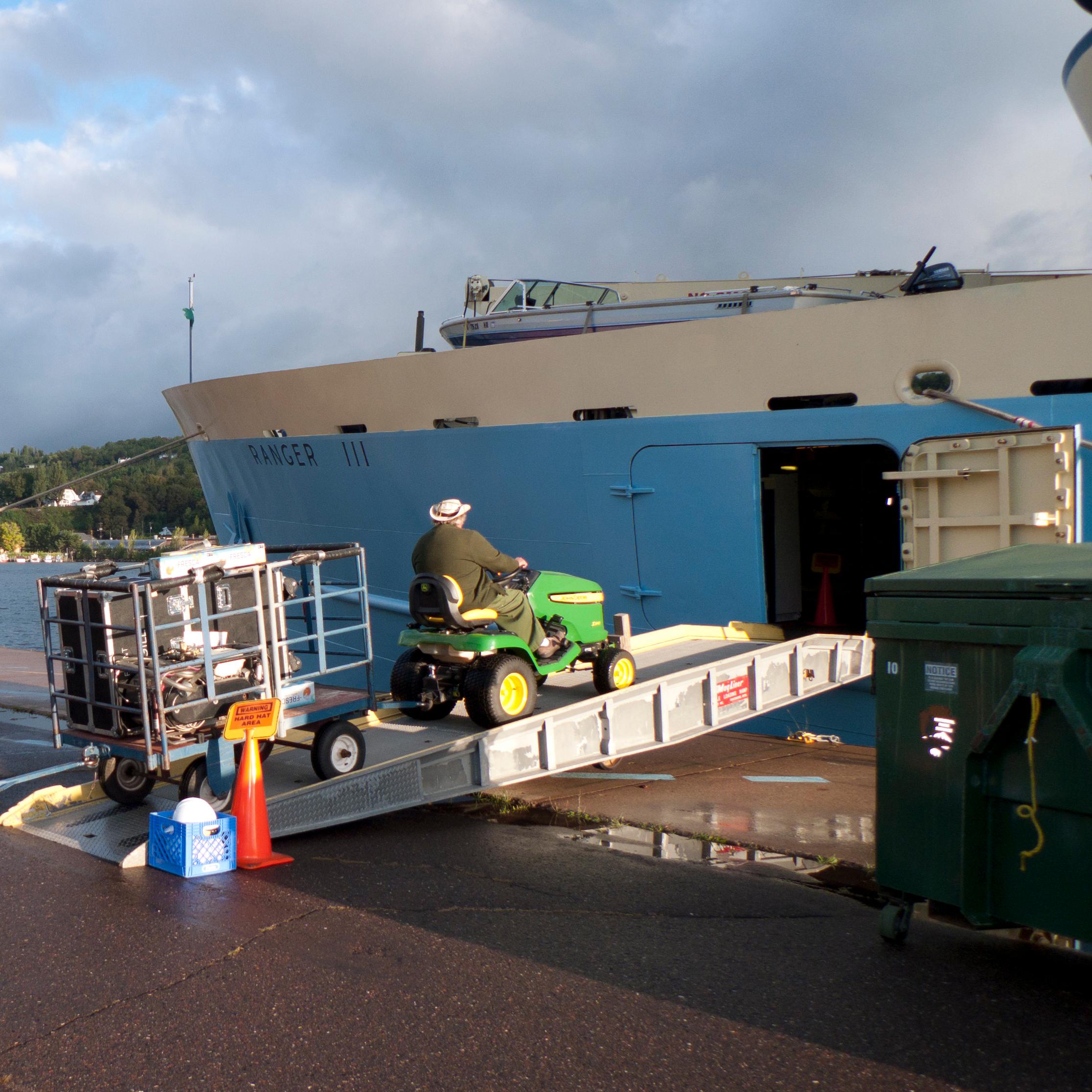 A tractor pulls two blue carts filled with baggage up a ramp into the cargo hold of RANGR III.