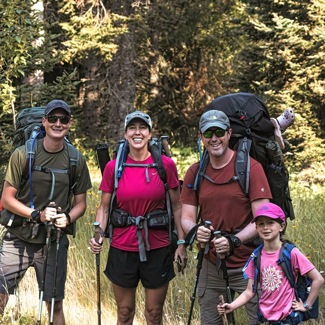 A group of three adults and one child poses for a photo with their backpacking packs on.
