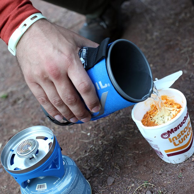 Human pours hot water from a Jet Boil stove into a Ramen Noodles container. 