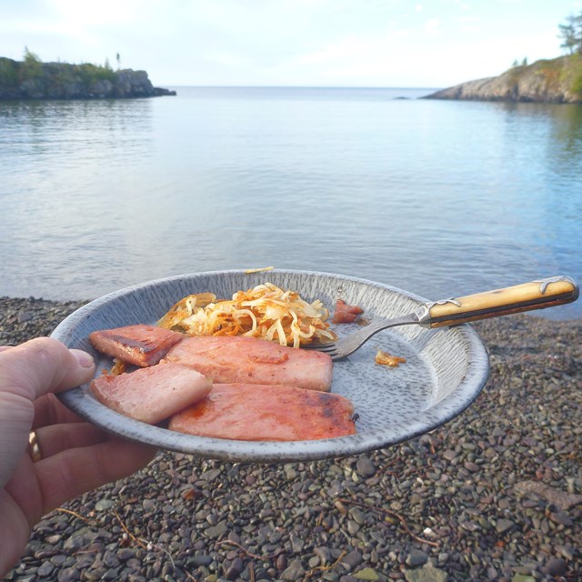 A camper holds a breakfast of hashbrowns and ham up to a lakeside background.