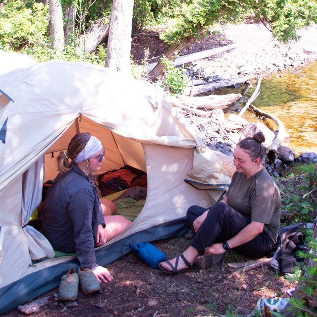 Two people talk while sitting on the ground near a tent.