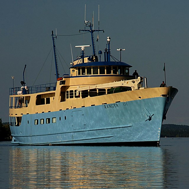 A large blue boat with white upper trim shines in the morning sun with Isle Royale in the background