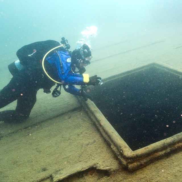 An underwater photo shows a scuba diver hovering above part of a shipwreck in clear water.