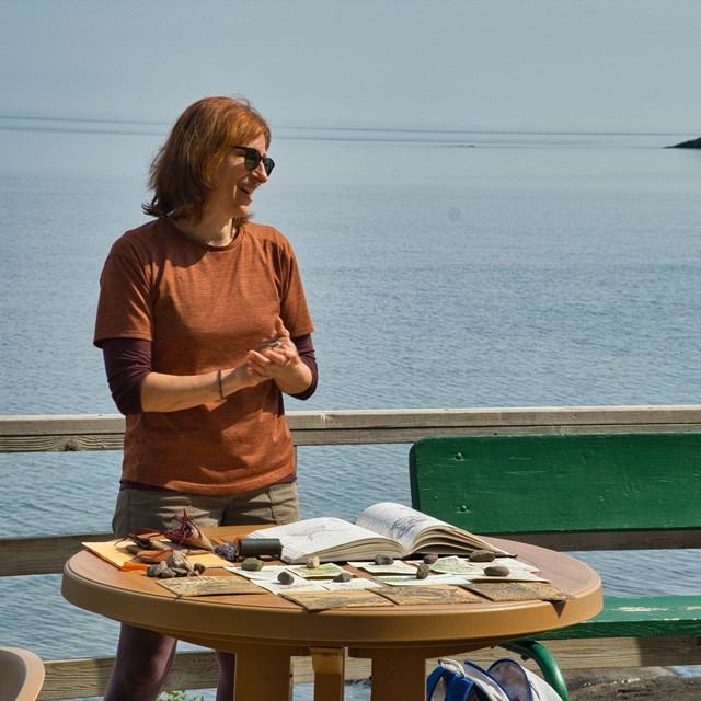 A smiling woman in an orange shirt stands at a round table displaying artwork.