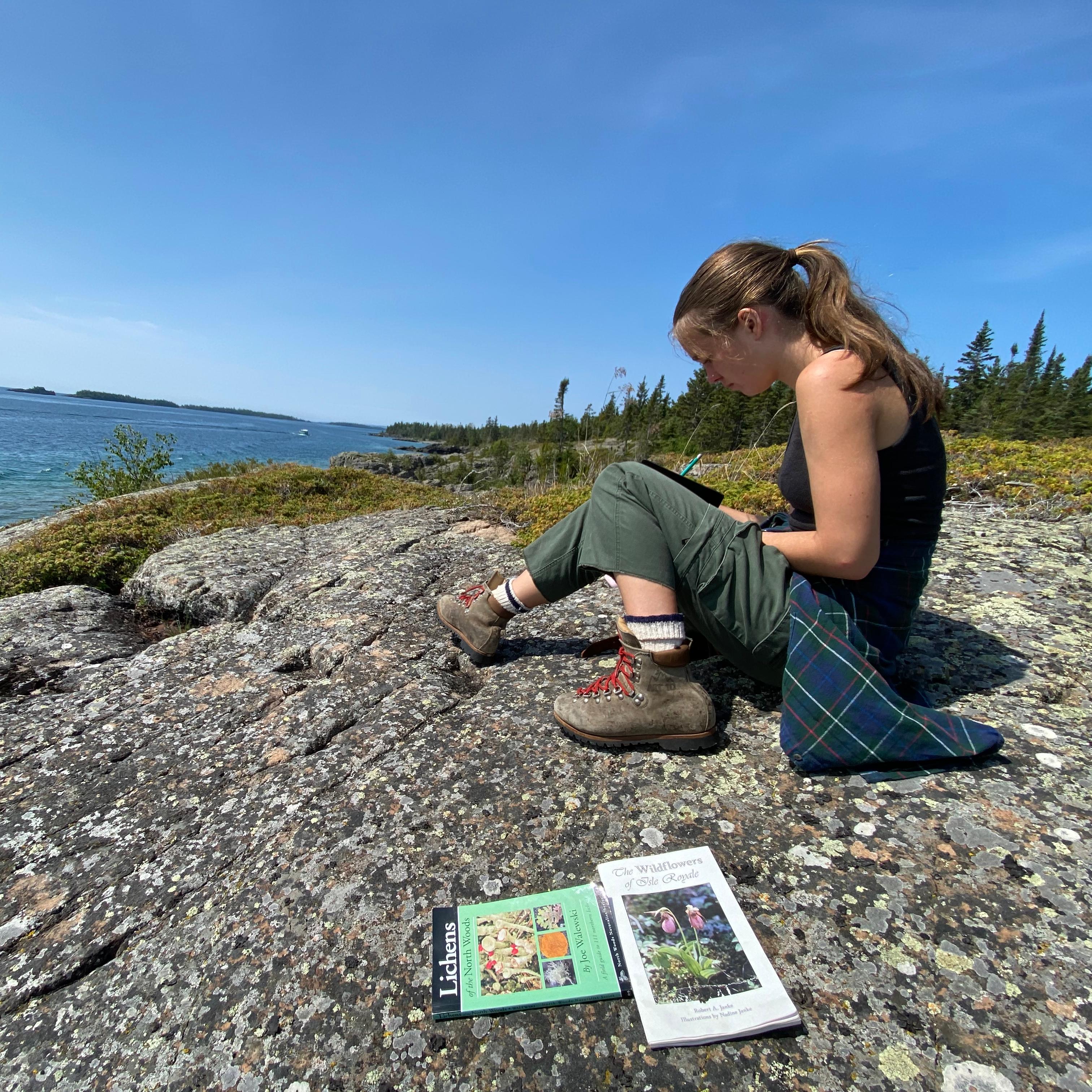 Teen Artist sitting on rocky Lake Superior shoreline, sketching with colored pencils.