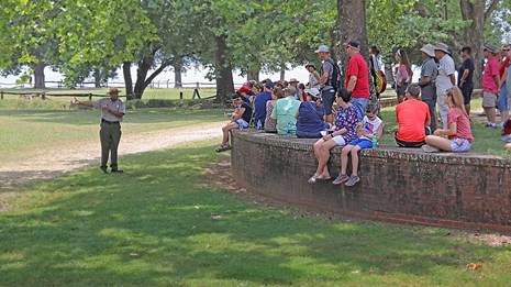 A park ranger speaks to a group of visitors as they sit along a curved brick wall.