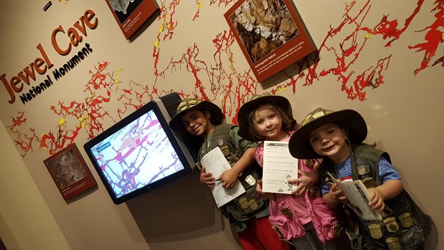 Three Junior Rangers stand in front of a large cave map, holding their activity booklets. 