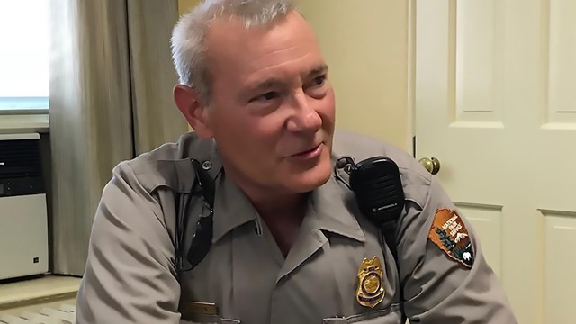 A park ranger sits at a table. His hands are on the table in front of him. He is smiling slightly.