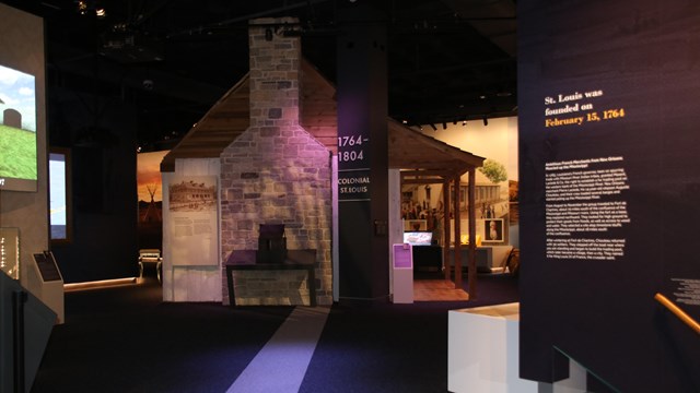 A dark museum room with text printed on the right wall. Farther back is a replica of a house.