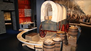 A model covered wagon, with some wooden barrels in the foreground and to the right side.