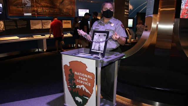 A ranger stands behind a podium. She holds a chain and a picture of the Arch.