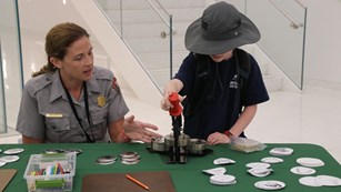 A child in a wide hat looks down at a button maker. A seated ranger points at the button maker
