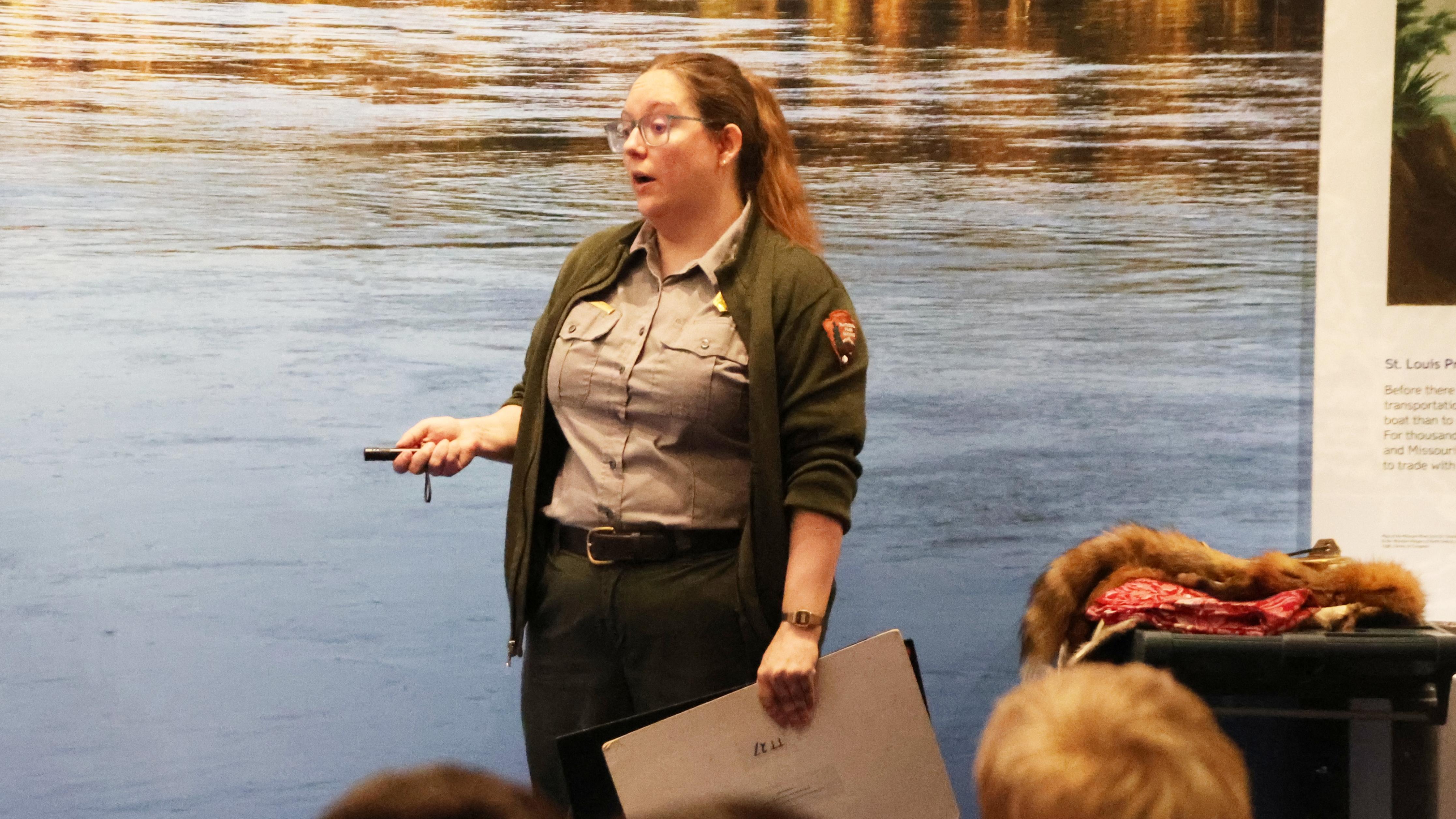 A ranger stands in front of a picture of a river. She holds an image and a pointer.