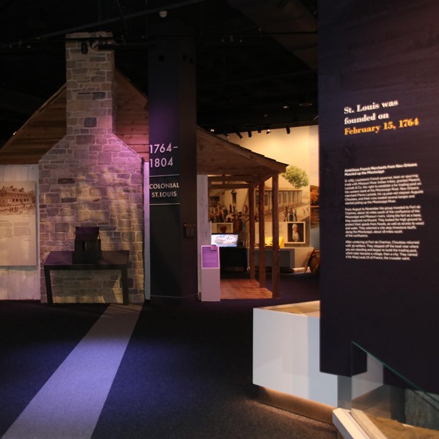 A dark museum room with text printed on the right wall. Farther back is a replica of a house.