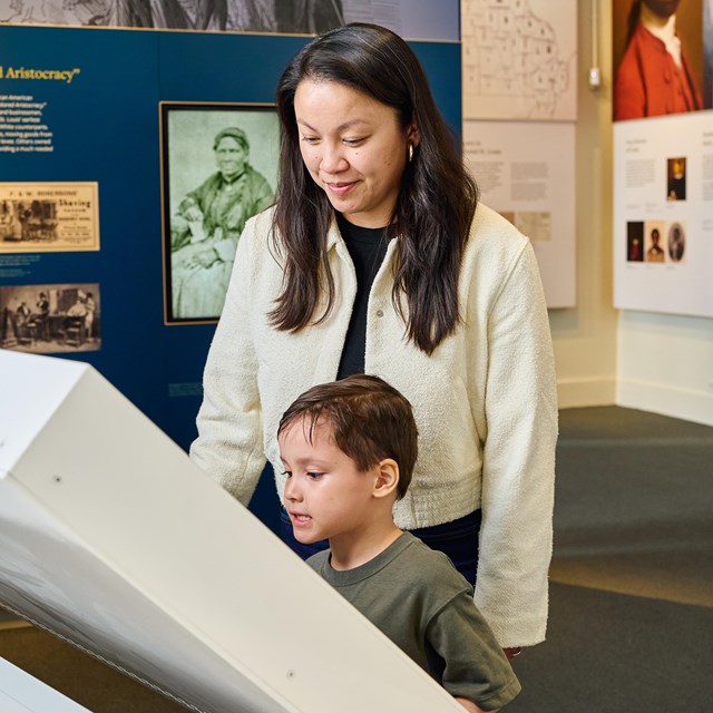 A child peers at a touchscreen in a museum while an adult looks on behind him