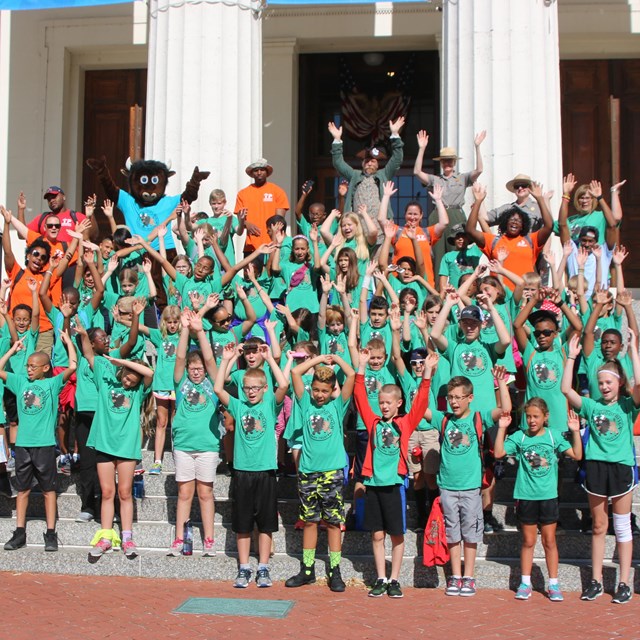 Large group of children and adults posing for a photo and raising hands while standing on steps.