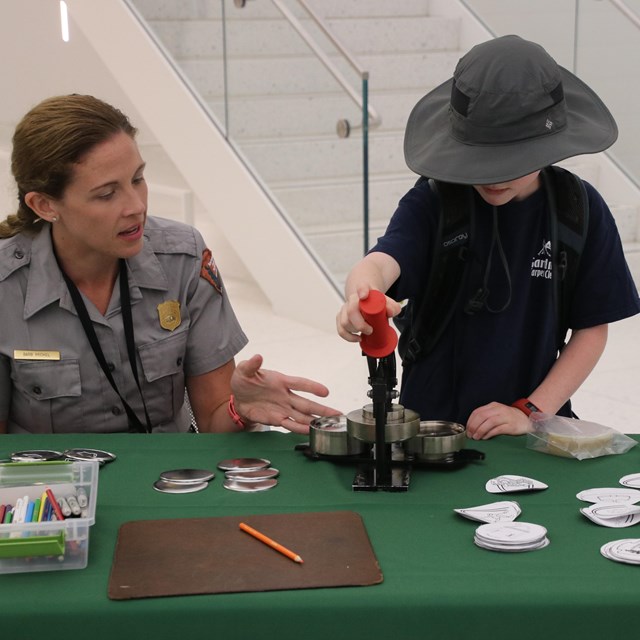 Park Ranger sitting in chair and working with child, who is using a button maker on a green table. 