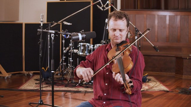 Man in a red button up shirt sits with a fiddle in his hands. A microphone is next to him.