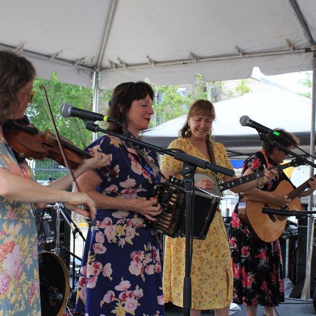 4 women stand on stage in front of microphones holding instruments.
