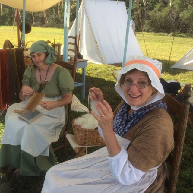 Two women wearing period clothing sit in chairs and hold cotton string and cording combs.