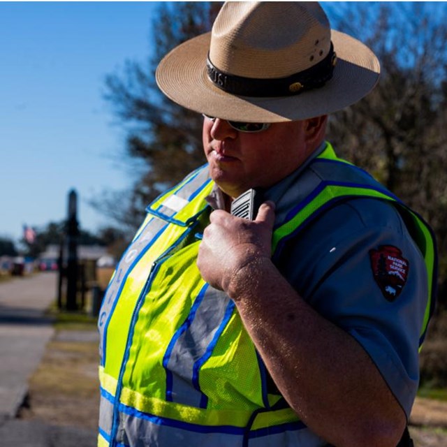 A law enforcement ranger in a flat hat & wearing a safety vest talks into a radio
