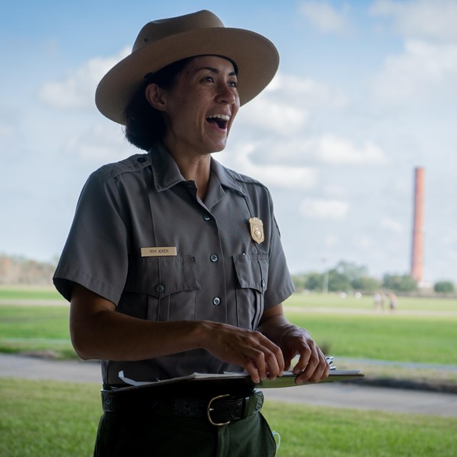 Park ranger stands smiling outside. She holds a clipboard.