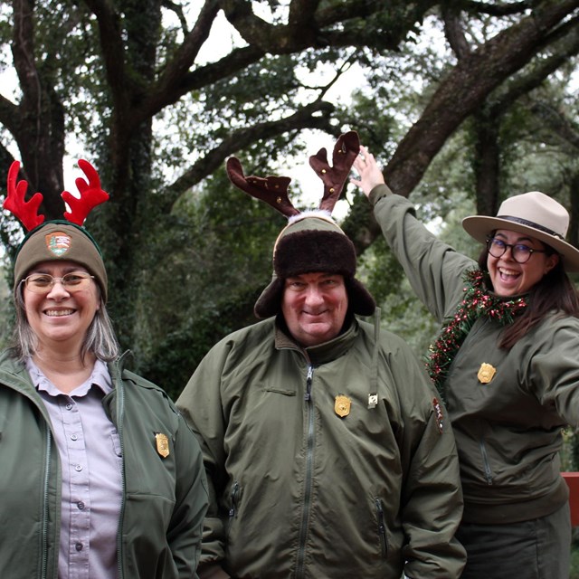 Three park rangers wear winter jackets and hats. 2 wear felt antlers, and 1 has a garland scarf.
