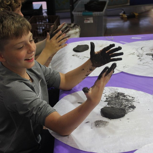 A kid smiles looking at muddy hands with a pile of clay in front of him.