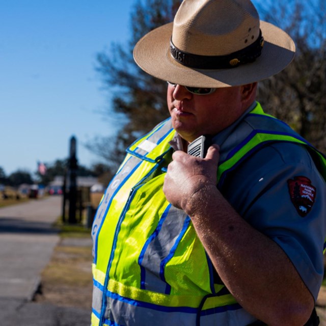 Law enforcement ranger stands in a safety vest and speaks into a radio.