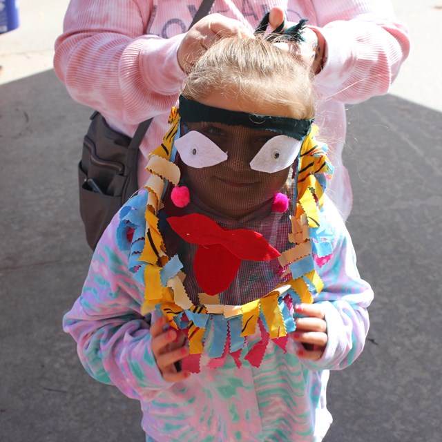 Outside daytime. Young girl smiles behind a mesh mask with felt on it.