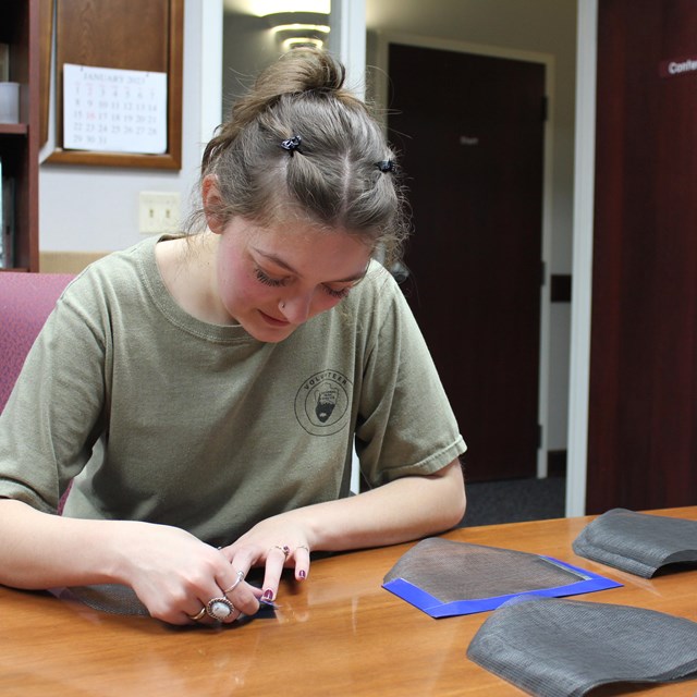 A young woman in a green t-shirt sits at a table with black mesh screens in piles to her left.