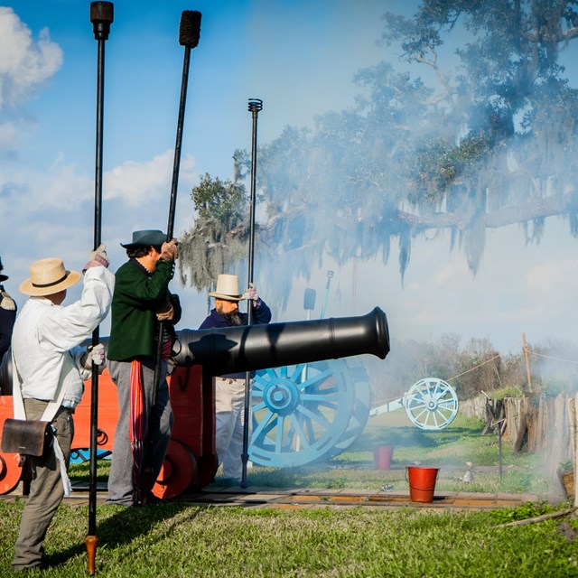 Outside daytime. Reenactors in period clothing stand next to a large black cannon. Smoke in air.