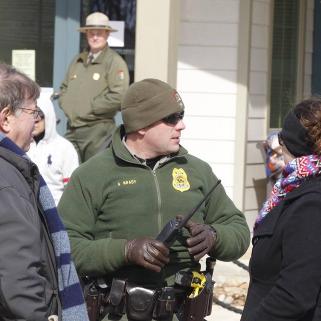 Law enforcement ranger stands outside next to two people.