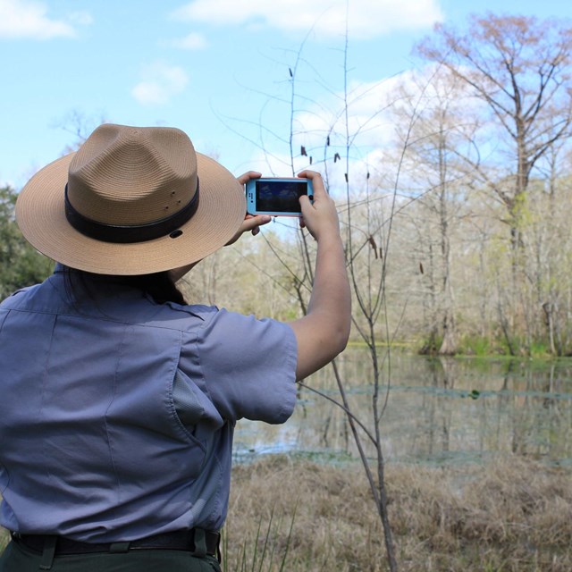 Park ranger stands outside next to a bayou waterway. She holds up a phone.