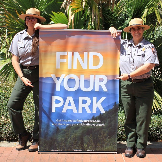 Two park rangers stand and point to a large sign that says 