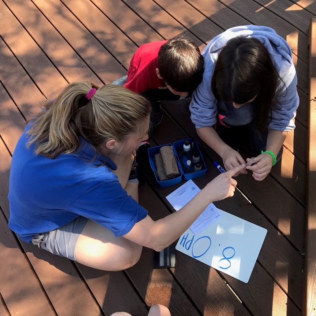 A young woman points to a test tube. Two kids look at the test tube. Small white board on ground.