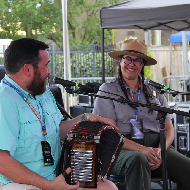A park ranger smiles at a man holding an accordion. Both sit in front of microphones.