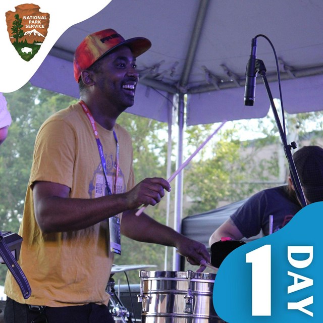 A man smiles and holds drum sticks. Text 