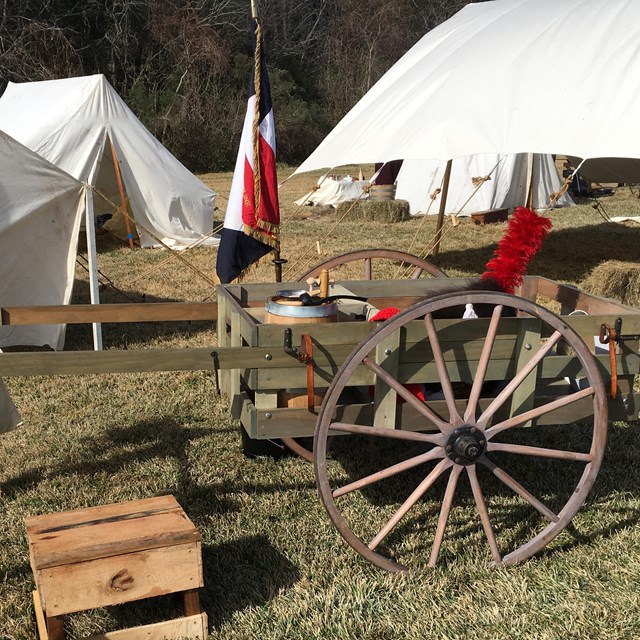 A replica of a hand cart from the War of 1812 sits in front of white canvass tents.